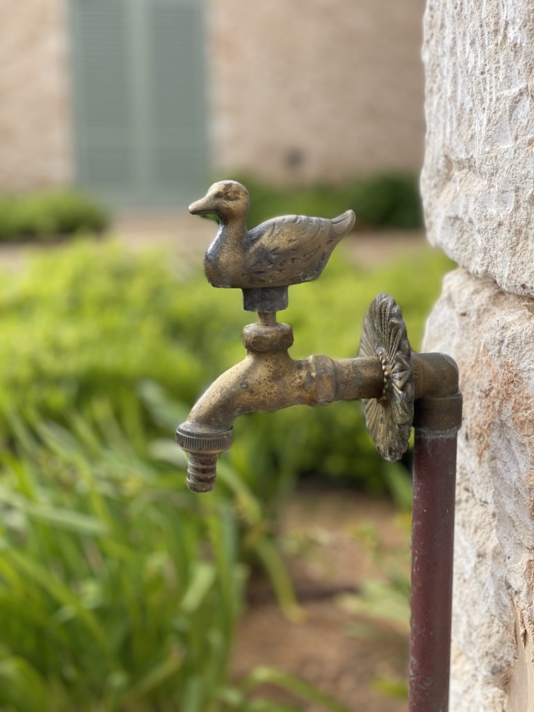 Brass garden tap with a cast duck on the handle, mounted on a natural stone pillar in a Mallorcan garden